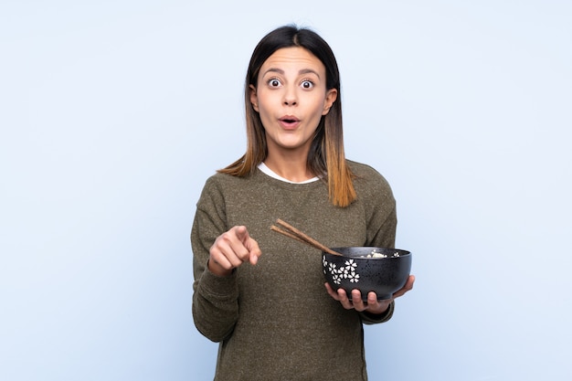 Mujer sobre pared azul aislada sorprendida y apuntando hacia delante mientras sostiene un tazón de fideos con palillos