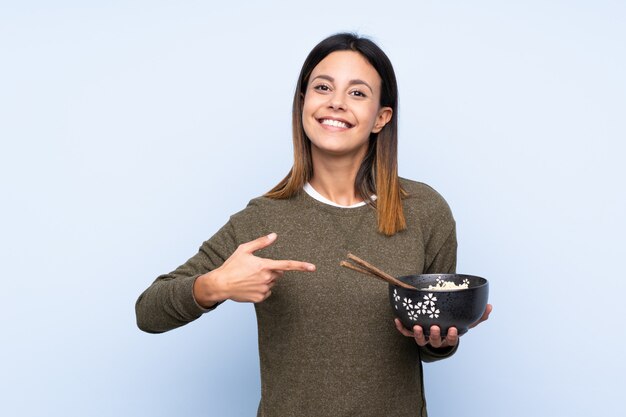 Mujer sobre pared azul y apuntando mientras sostiene un tazón de fideos con palillos