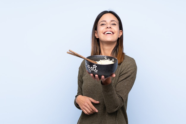 Mujer sobre pared azul con expresión feliz mientras sostiene un tazón de fideos con palillos