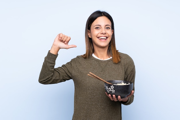 Mujer sobre pared azul orgullosa y satisfecha mientras sostiene un plato de fideos con palillos