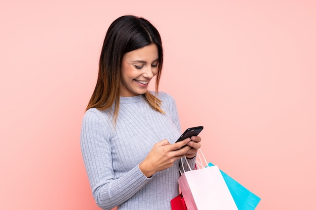 Mujer sobre pared rosa aislada sosteniendo bolsas de compras y escribiendo un mensaje con su teléfono celular a un amigo