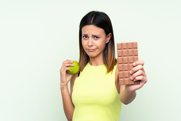 Mujer sobre pared verde aislada tomando una tableta de chocolate en una mano y una manzana en la otra