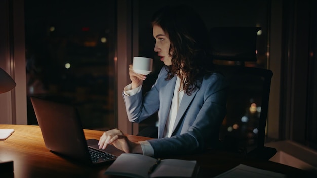 Mujer sobrecargada de trabajo bebiendo café escribiendo en la computadora portátil por la noche