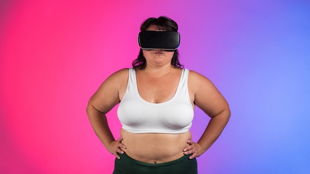 Mujer con sobrepeso en chándal y auriculares VR posando en un estudio con fondo azul y rojo