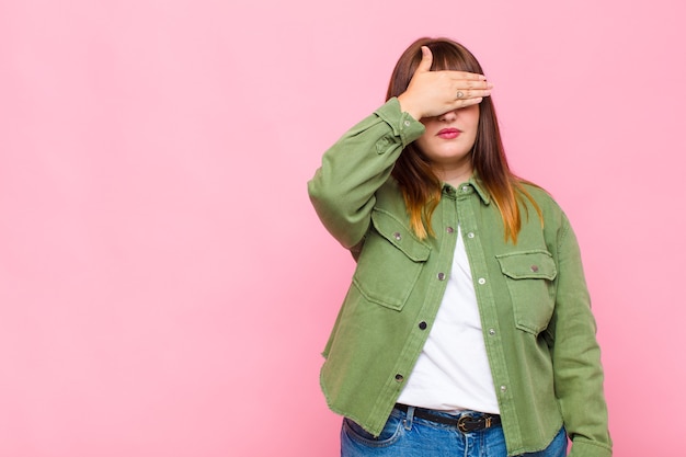 Mujer con sobrepeso cubriendo los ojos con una mano sintiéndose asustada o ansiosa, preguntándose o esperando ciegamente una sorpresa