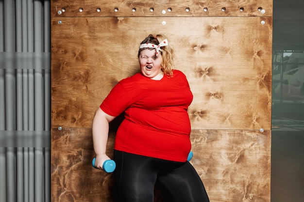 Mujer con sobrepeso divertida vestida con ropa deportiva y con un vendaje en la cabeza se encuentra con pesas en el fondo de una pared de madera en el gimnasio