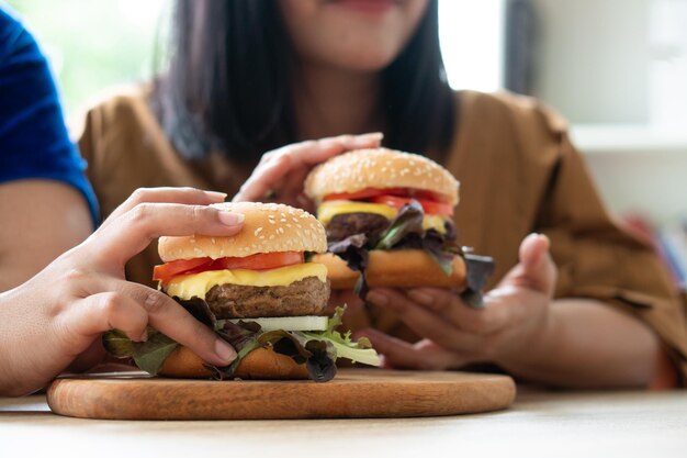 Mujer con sobrepeso hambrienta sosteniendo una hamburguesa en una placa de madera, durante el trabajo desde casa, problema de aumento de peso. Concepto de trastorno por atracón (BED).