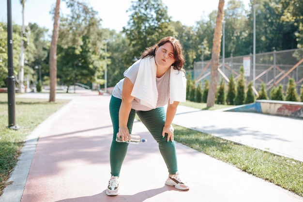 Una mujer con sobrepeso en ropa deportiva hace una pausa afuera con su botella de agua y su estilo de vida saludable