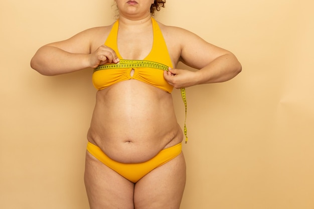 Foto mujer con sobrepeso en traje de baño amarillo mide la circunferencia del busto con cinta métrica fondo beige adelgazamiento y control del cuerpo lucha contra la obesidad personas de talla grande y concepto positivo del cuerpo