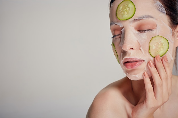 Foto mujer sofisticada con una máscara hidratante con un pepino fresco en la cara en la serie con los ojos cerrados y la mano cerca de la cara