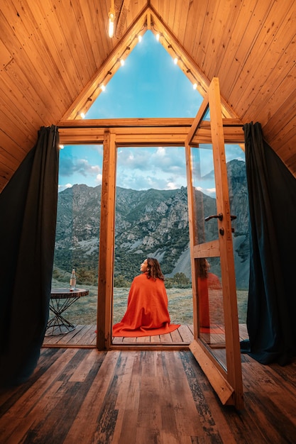 Foto mujer sola en el porche de una pequeña casa de madera en las montañas el concepto de glamping y vacaciones idílicas