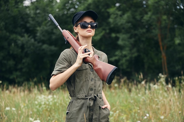 Mujer soldado con un arma en gafas oscuras cazando en el fondo del bosque de gorra negra