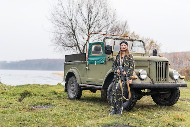 Foto mujer soldado con rifle posando cerca de coche militar