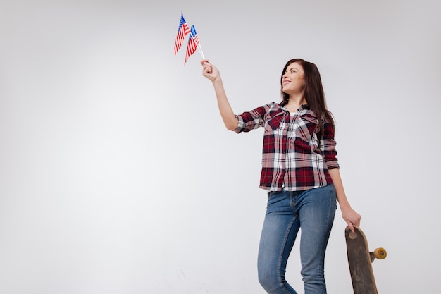 Foto mujer soleada alegre delgada sonriendo y celebrando el día de la independencia americana mientras está de pie contra la pared blanca y sosteniendo la bandera americana y el monopatín