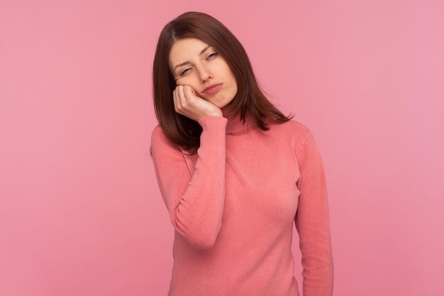 Mujer solitaria y cansada con suéter rosa inclinada cabeza a mano, mirando con expresión aburrida, exhausta por el exceso de trabajo. Disparo de estudio interior aislado sobre fondo rosa