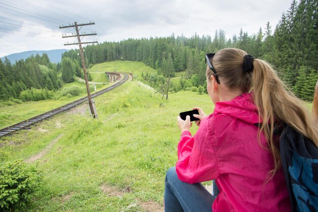 Mujer soltera sentada cerca del ferrocarril y mirando al teléfono inteligente