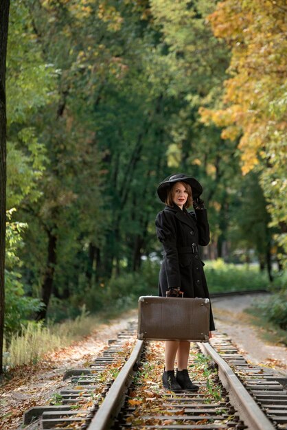 Mujer con sombrero y abrigo sostiene maleta retro bosque de otoño en vía férrea Dama con ropa de estilo 3040Marco vertical