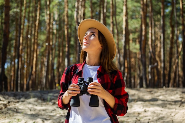 Mujer con sombrero y camisa a cuadros roja con binoculares en el fondo del bosque.