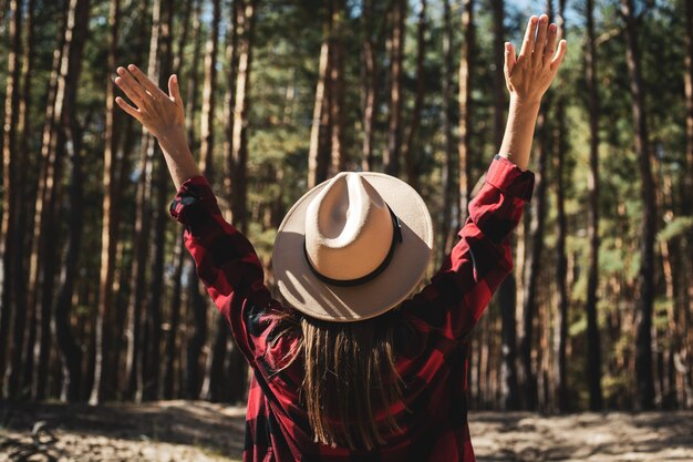 Foto mujer con sombrero y camisa a cuadros roja en el bosque.