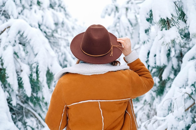 Foto mujer con sombrero y chaqueta abrigada de pie sobre un fondo de bosque nevado vista desde atrás