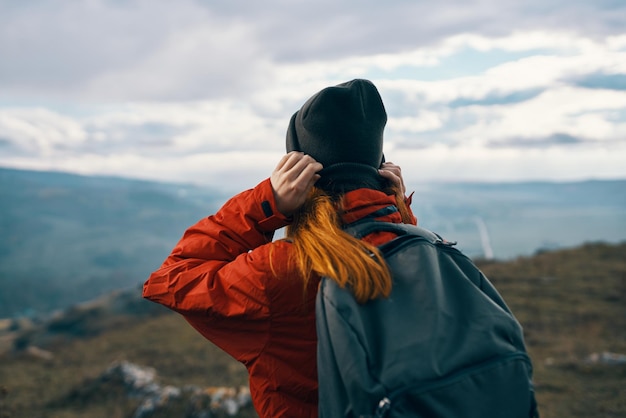 Mujer con un sombrero de chaqueta y una mochila mira las montañas en la distancia en la naturaleza