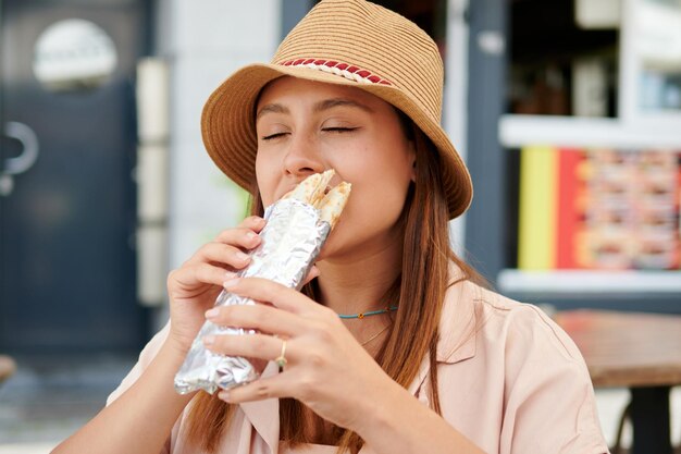 Una mujer con sombrero comiendo un kebab en un café de la calle