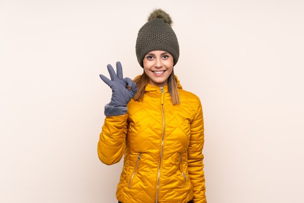 Mujer con sombrero de invierno sobre la pared mostrando un signo bien con los dedos