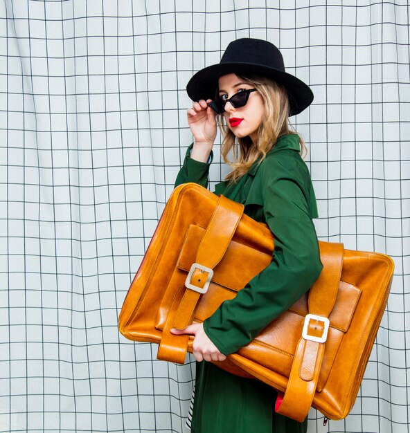 Mujer con sombrero y manto verde estilo años 90 con maleta de viaje.