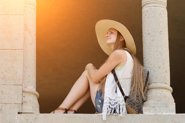 mujer con sombrero y mochila está sentada cerca de la columna.