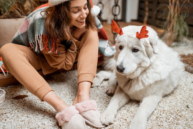 Mujer con sombrero de navidad celebrando con su lindo perro unas vacaciones de año nuevo, sentados juntos en la terraza bellamente decorada en casa