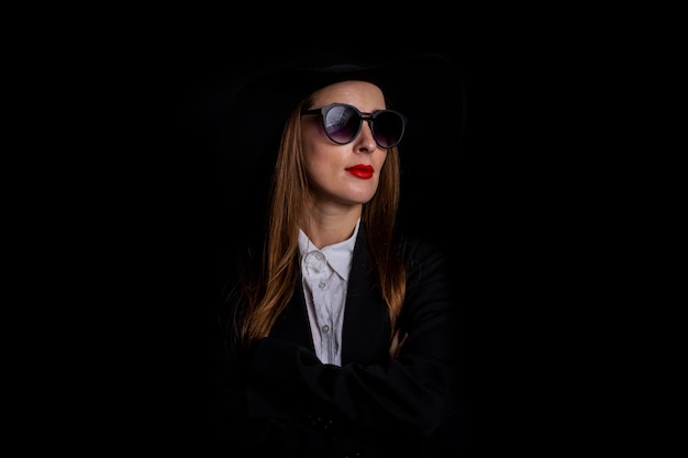 Mujer con sombrero negro, gafas y chaqueta sobre un negro.