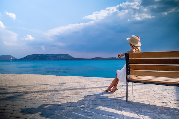 Mujer con sombrero de paja para el sol sentado en un banco de madera en el puerto deportivo de Ermioni, Grecia.