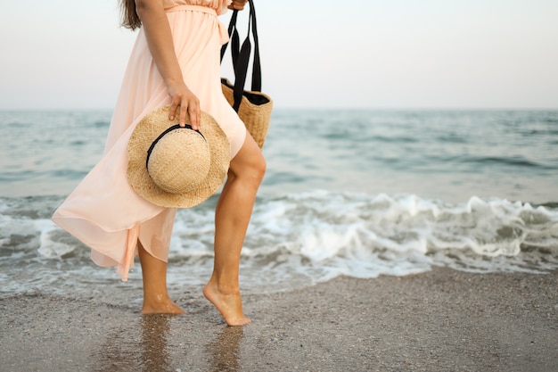 Mujer con sombrero de paja y vestido rosa en una playa tropical con bolso marrón.