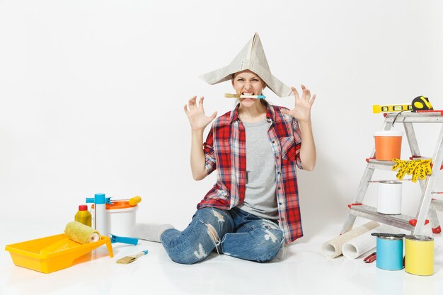 Mujer con sombrero de periódico sentada en el suelo con cepillo en la boca, instrumentos para apartamento de renovación aislado sobre fondo blanco. Papel pintado, accesorios para pegar, herramientas de pintura. Reparar el concepto de hogar.