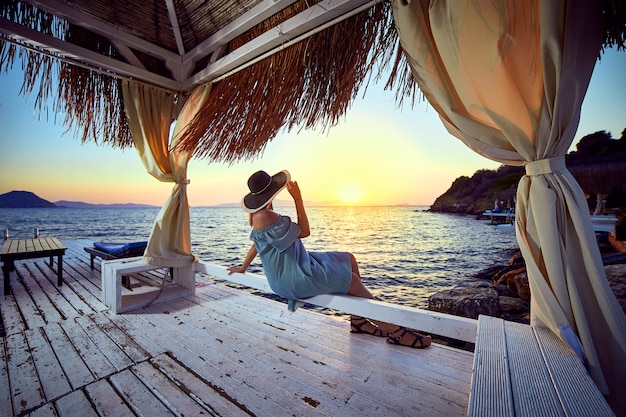 Mujer con sombrero relajante junto al mar en un lujoso complejo hotelero frente al mar al atardecer disfrutando de unas vacaciones perfectas en Bodrum, Turquía. Concepto de viaje de verano de paisaje marino al aire libre