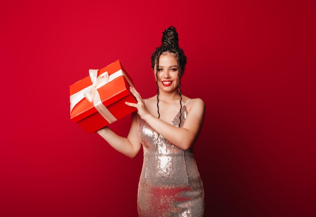 Una mujer con un sombrero rojo de Navidad con rastas sostiene un regalo sobre un fondo rojo Aísla a una mujer con un vestido elegante año nuevo navidad