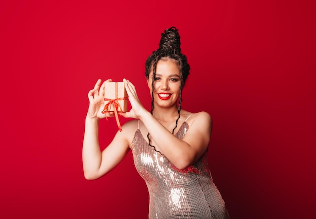 Una mujer con un sombrero rojo de Navidad con rastas sostiene un regalo sobre un fondo rojo Aísla a una mujer con un vestido elegante año nuevo navidad