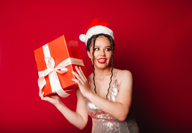 Una mujer con un sombrero rojo de Navidad con rastas sostiene un regalo sobre un fondo rojo Aísla a una mujer con un vestido elegante año nuevo navidad