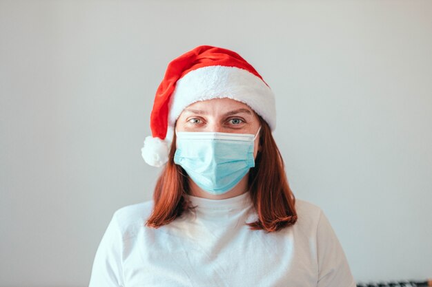 Mujer con sombrero de santa y máscara médica protectora sobre fondo de pared gris. Año nuevo en cuarentena