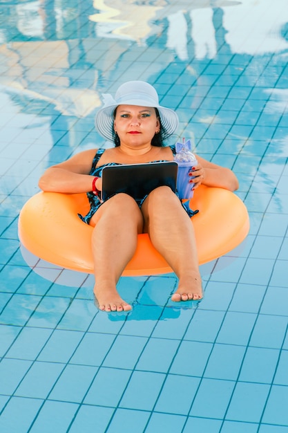 Mujer con sombrero para el sol y traje de baño en la piscina en un círculo de natación con una computadora portátil en su regazo.