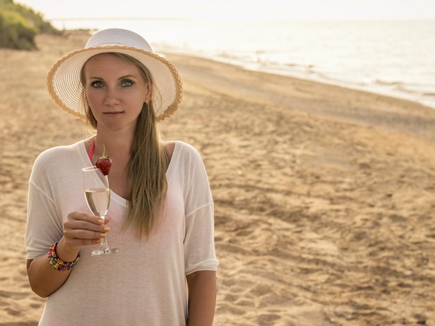 Mujer con sombrero sosteniendo una copa de champán y fresas en una playa de arena.