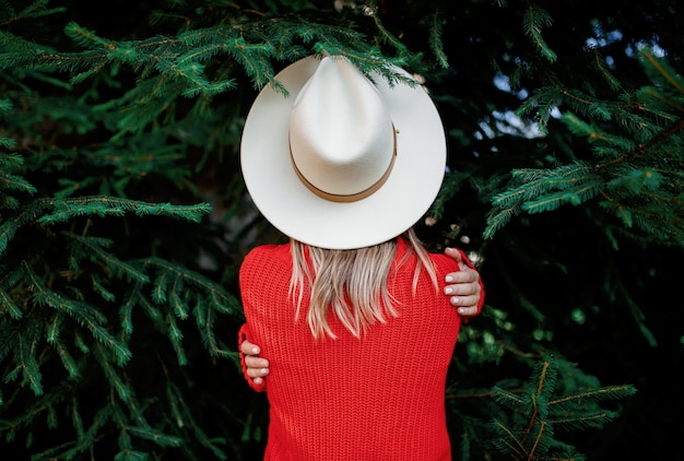 Mujer con sombrero y suéter rojo permanecer cerca de pino.