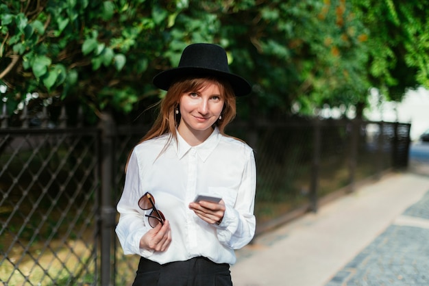 Mujer con sombrero tiene gafas y un teléfono inteligente y mira a la cámara