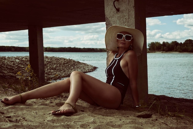 Mujer con sombrero de traje de baño y gafas de sol se sienta en la arena en verano en la orilla del río en un muelle de hormigón