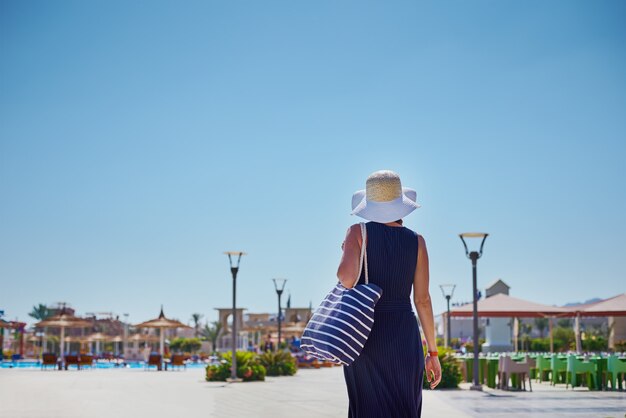 Mujer con sombrero y vestido azul salir a caminar en el hotel resort cerca de la piscina con bolsa de playa