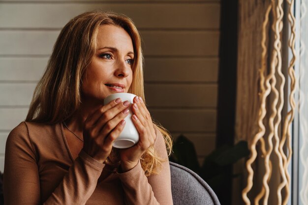 Mujer soñadora de mediana edad en la sala de estar con una taza de té o café mirando hacia otro lado