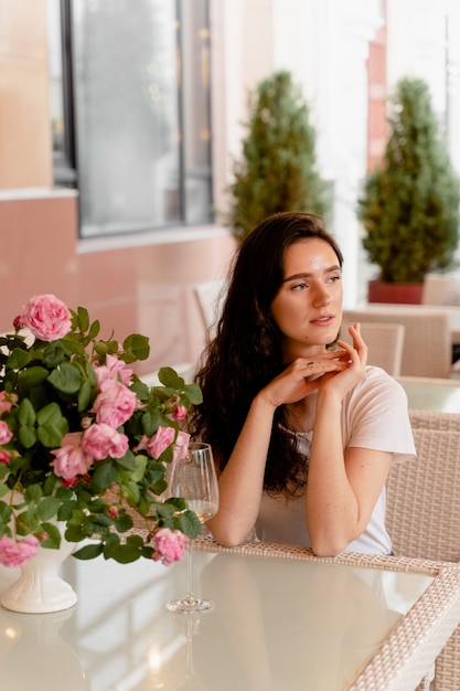 Mujer soñando con copa de vino blanco alcohólico en café en terraza de verano y ramo de rosas sobre la mesa.