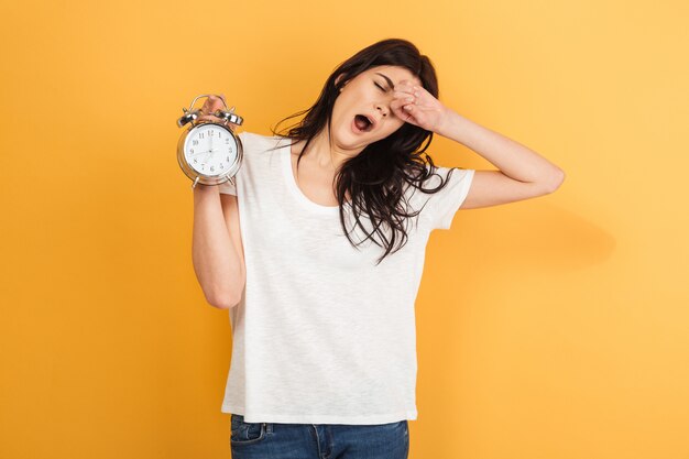 Mujer soñolienta aislada sosteniendo el despertador bostezando.