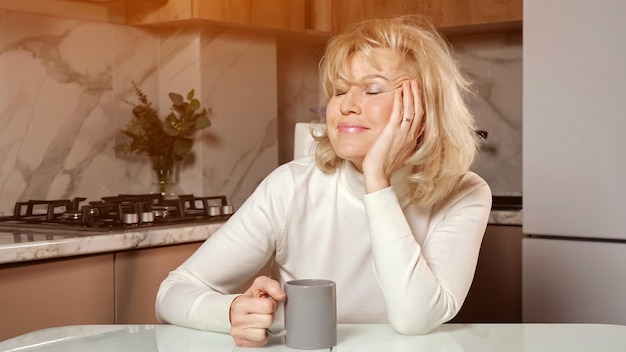 Una mujer soñolienta de mediana edad bebe café sentada en la cocina