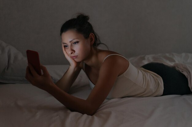 Mujer soñolienta mirando su teléfono inteligente en la cama por la noche. Dependencia y uso excesivo del teléfono. Concepto de adicto a los gadgets.